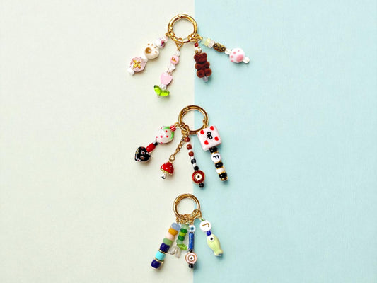 May include: Three keychains with gold rings. Each keychain has colorful beads and charms. One has flower, heart, and bear charms. Another has a dice and letter charms. The last has a fish and letter charms. The background is split between light blue and white.