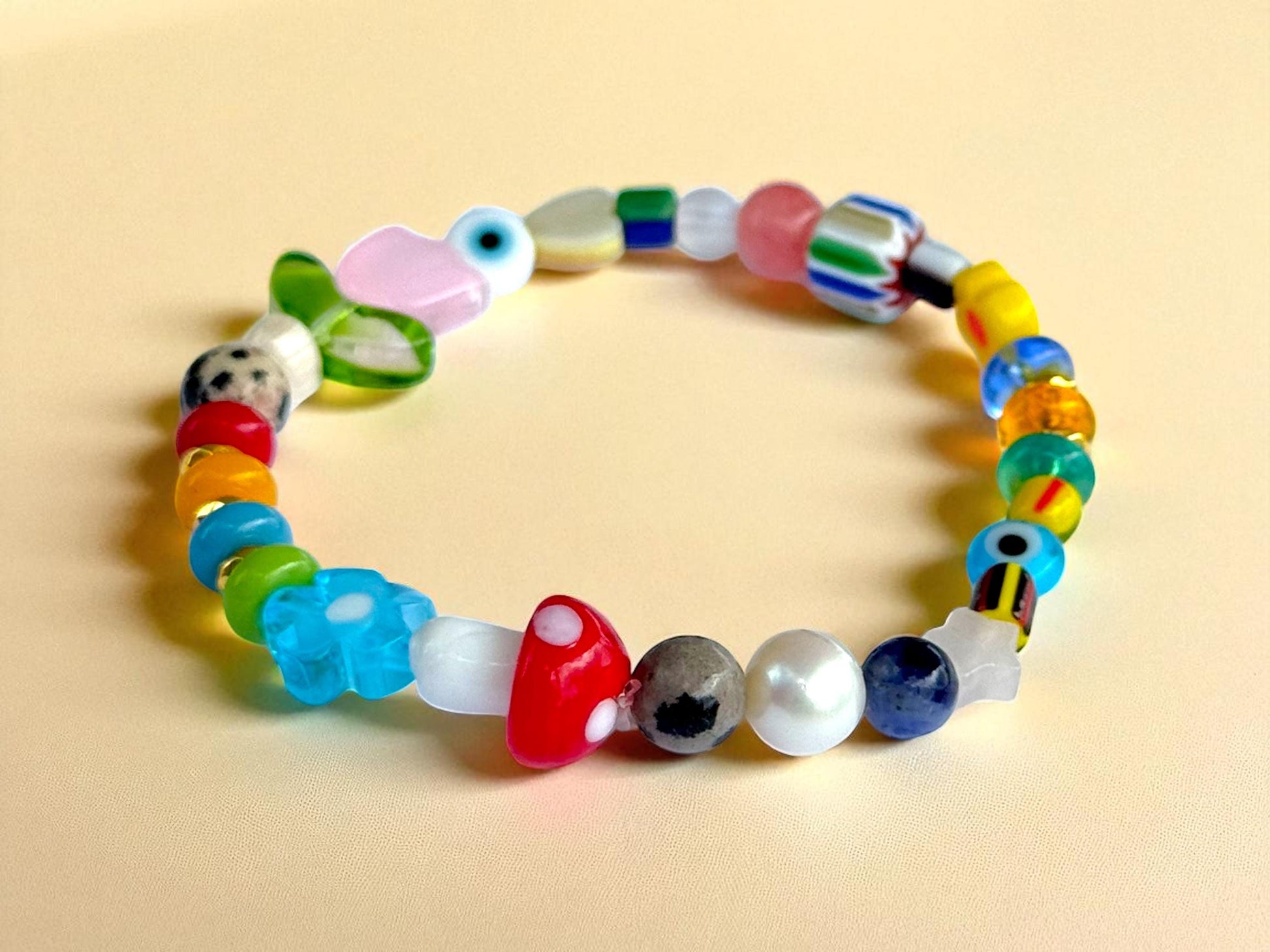 May include: A colorful beaded bracelet with a variety of shapes and colors. The bracelet features round, oval, and flower-shaped beads in shades of red, blue, green, yellow, and pink. Some beads have patterns, including a mushroom and a duck. The bracelet is on a light yellow surface.