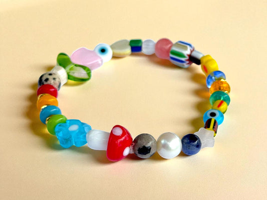 May include: A colorful beaded bracelet with a variety of shapes and colors. The bracelet features round, oval, and flower-shaped beads in shades of red, blue, green, yellow, and pink. Some beads have patterns, including a mushroom and a duck. The bracelet is on a light yellow surface.