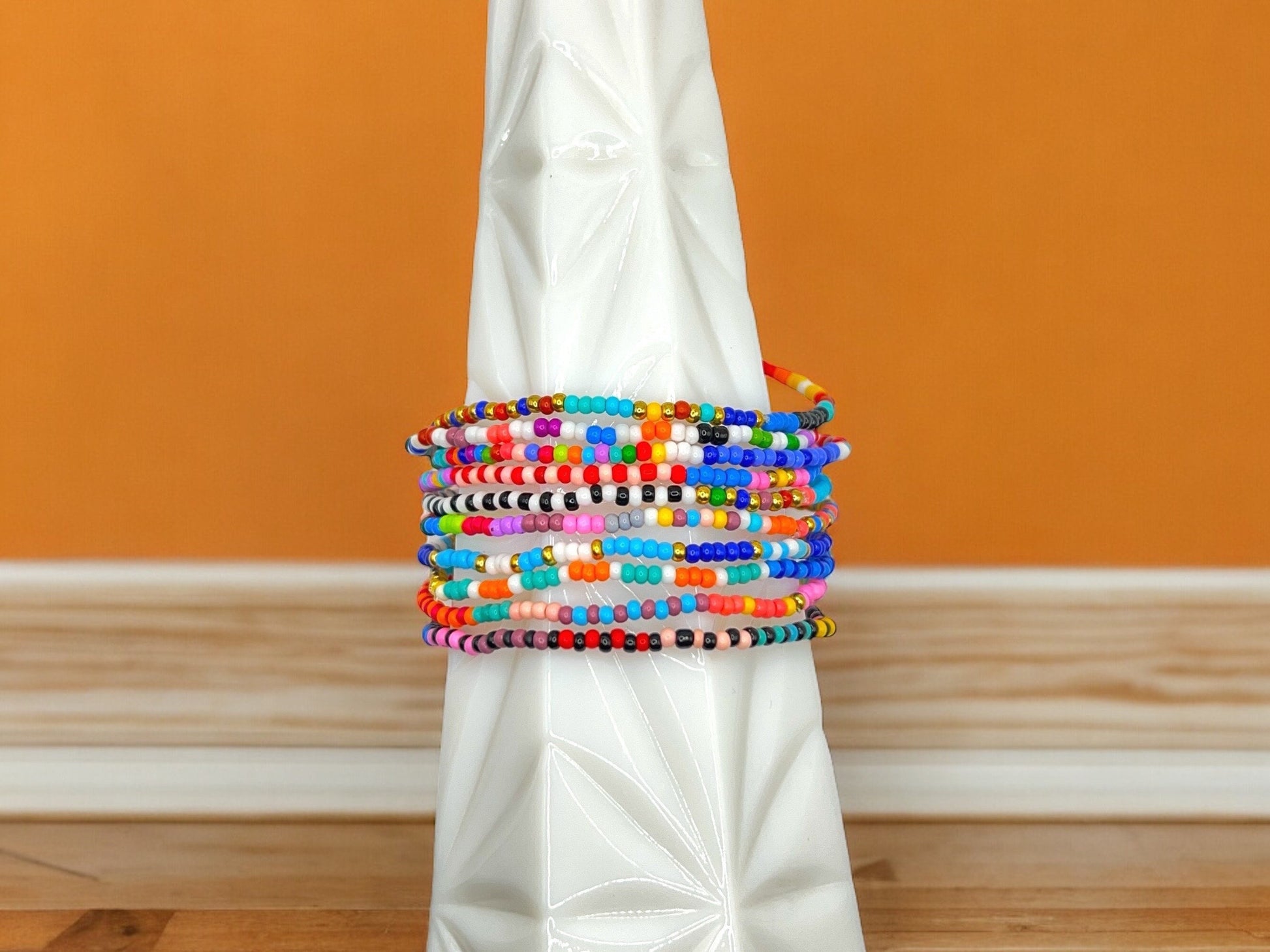 May include: A stack of colorful beaded bracelets in shades of blue, red, orange, yellow, green, pink, white, black, and gray. The bracelets are stacked on top of each other on a white ceramic vase.
